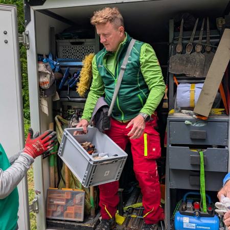 Ein Wegearbeiter gibt Werkzeug aus dem Wegebauanhänger aus - TRAILS Project